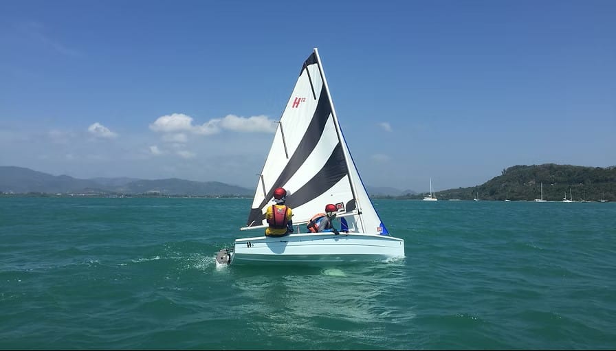 Sailors training aboard a Hartley 12s dinghy with black and white sail on tropical waters in Thailand.