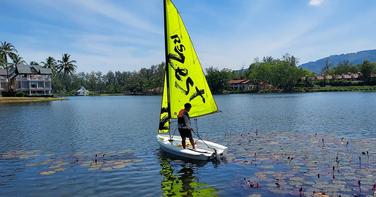 Sailor handling a bright RS Zest dinghy on calm water during practical sailing training in Thailand.