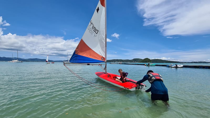 Instructor assisting a student with a Topper sailing dinghy in shallow water during a Java Yachting course in Phuket.